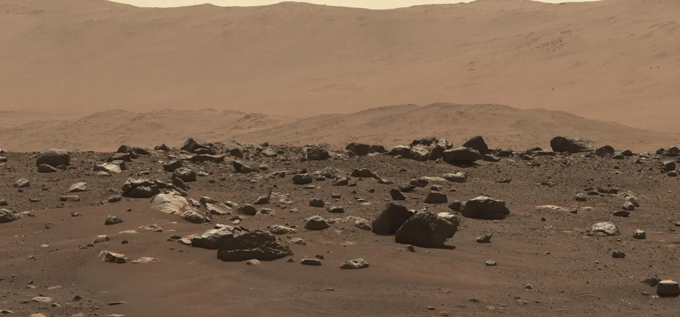 The floor of Jezero Crater on Mars: rocks in the foreground and the cliffs of an ancient river delta behind, under a dusty sky.